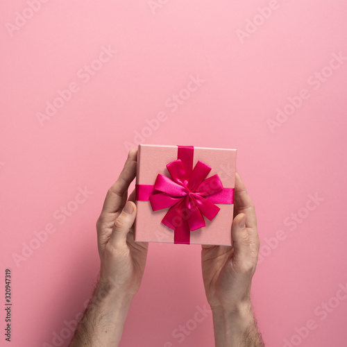 Fototapeta Naklejka Na Ścianę i Meble -  Valentine's Day celebration concept. A nice gift from a loved one. Box with a bow in male hands on a delicate pink background. Copy space. Flat lay. Square.