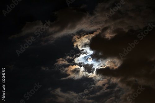 Full moon shining glowing light through the darkness of cloudy night sky; long exposure for beautiful nature background.