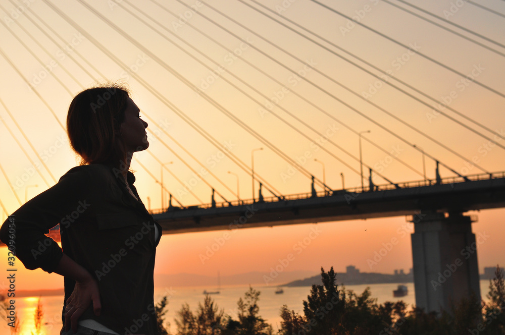 Fototapeta premium the silhouette of a young beautiful girl with her hands on her waist looks at a large cable-stayed bridge on the background of the sunset, a moment of enjoying life in a big city, matte mood toning