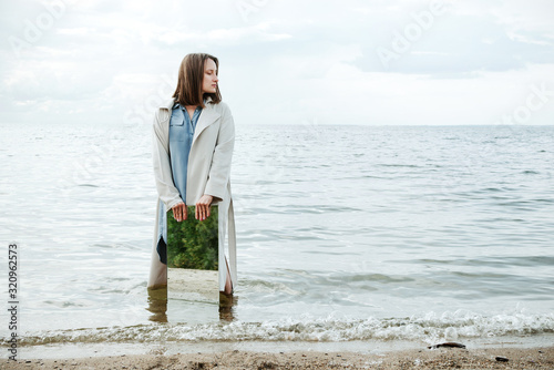 Closeup of woman standind on a shore in blue dress holding a mirror with beach reflecting in it, selective © lavrenkova