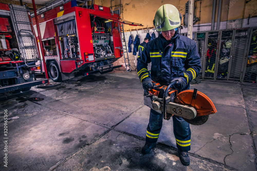 A fireman with uniform and helmet holding a chainsaw with fire truck in the background A fireman with uniform and helmet holding a chainsaw with fire truck in the background
