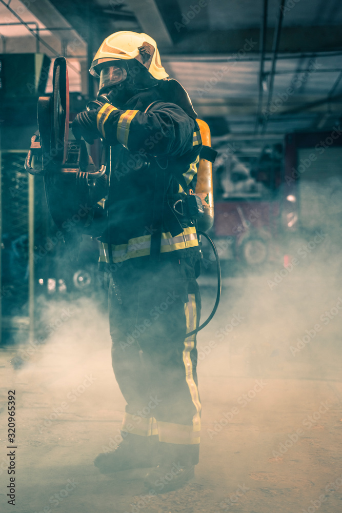 Portrait of a firefighter wearing full protective equipment posing with a chainsaw on his shoulder. Dark background with smoke and blue light.