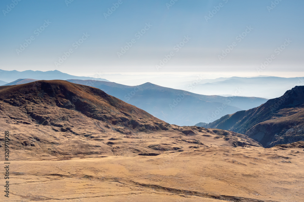 View from above of a panoramic mountain landscape at аutumn, alternating mountain ranges, covered in fog and clear blue sky in the background, Rila National Park, Bulgaria.