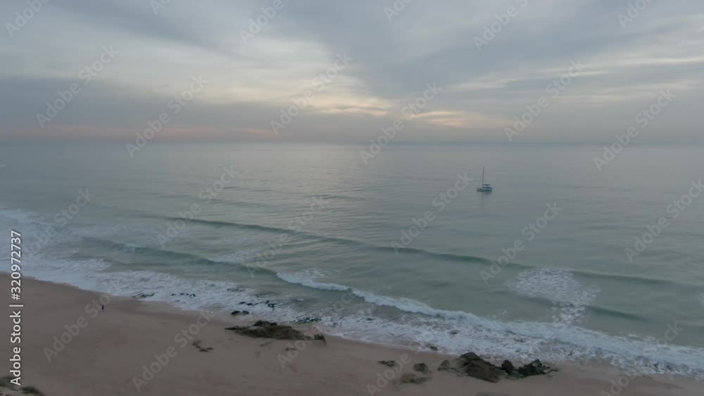 Aerial view of Person standing on empty Beach looking at sailboat at sunset
