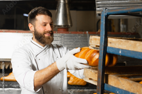 A baker stacks fresh hot bread in a rack against the background of an industrial oven in a bakery. Automatic bread making line