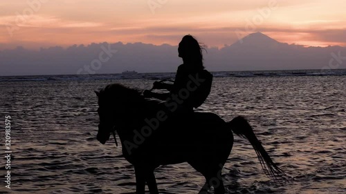 Silhouette slow motion shot of man riding a horse during sunset at the beach of Gili Trawangan, Indonesia. Ocean and active volcano Mount (Gunung) Agung in Bali covered in clouds in the background.