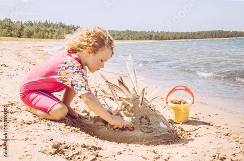 5 year old girl enjoy play in sand wearing pink long UV protective swimsuit, protecting light sensitive skin from sunburn. Kid building sandcastle in summer empty beautiful beach in Hiiumaa island. photo