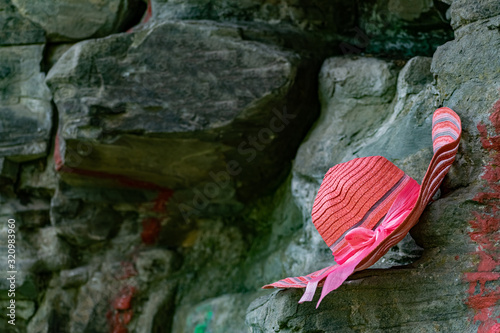 A bright and rich pink hat with a stripe pattern and a matching satin ribbon bow hangs on large stone rocks in a darkened place with sunlight.