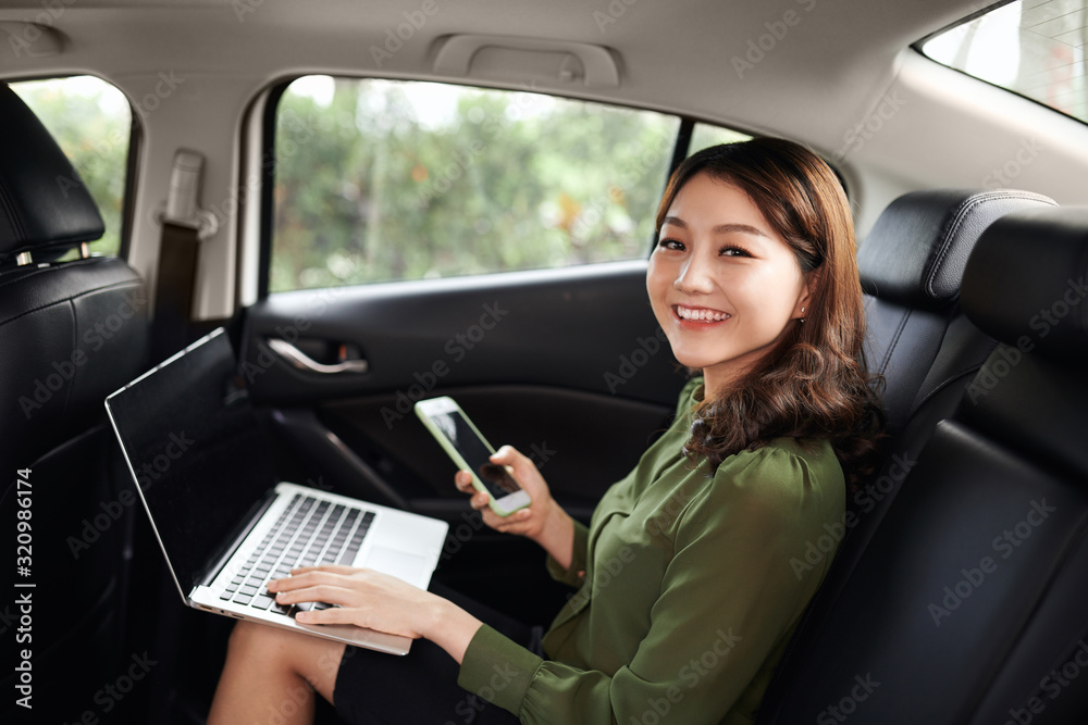 Photo of young asian woman talking on the phone while working with laptop computer in the car