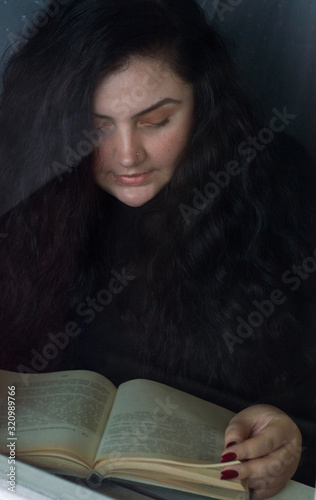 portrait of young woman plus size with wealth of brown hair, she is reading a book next to the window