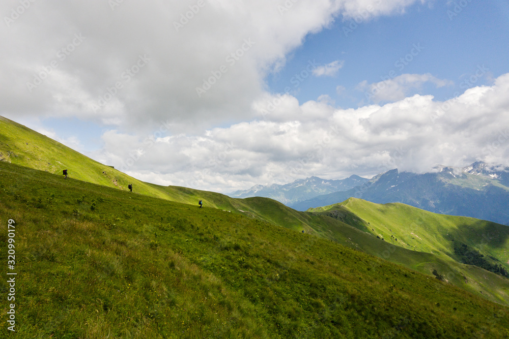 Hiker with a backpack goes on the grassy slope on a background of mountains. Back view. Beautiful sunny day in Arkhyz. Concept of healthy lifestyle, trekking activity, hiking adventure