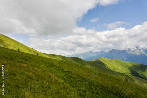 Hiker with a backpack goes on the grassy slope on a background of mountains. Back view. Beautiful sunny day in Arkhyz. Concept of healthy lifestyle  trekking activity  hiking adventure