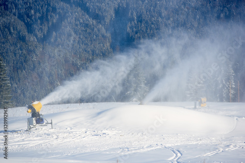 Electric snow gun machine pouring with white snow on mountain resort. Artificial snowfall in sunny day. Preparation for beginning ski season. Concept of combination of technology and nature.