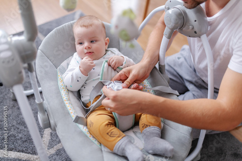 Valokuva Handsome smiling young caucasian dad putting his adorable 6 months old son in baby rocker chair