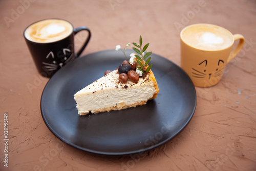 Two cups with a coffee stand near a black plate on which lies a piece of cake on which there are nuts and fruits