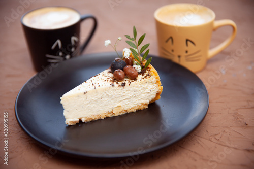 Two cups with a coffee stand near a black plate on which lies a piece of cake on which there are nuts and fruits