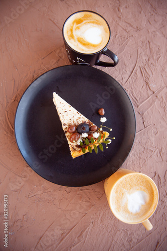 Two cups with a coffee stand near a black plate on which lies a piece of cake on which there are nuts and fruits