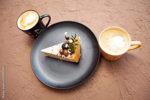 Two cups with a coffee stand near a black plate on which lies a piece of cake on which there are nuts and fruits