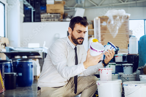 Young handsome caucasian graphic engineer in shirt and tie crouching next to buckets with colors  holding cans and looking at it. Printing shop interior.