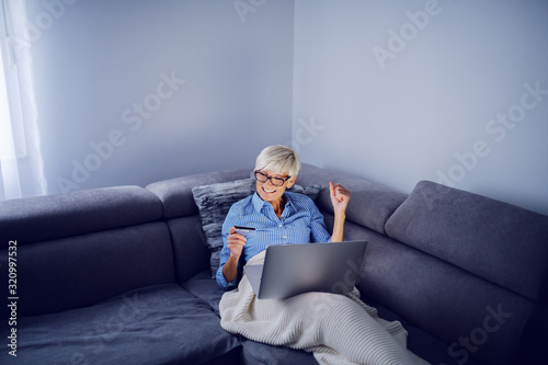 Cheerful charming caucasian senior blond woman with short hair and with eyeglasses sitting on sofa in living room, holding laptop in lap and using credit card for online shopping. © dusanpetkovic1