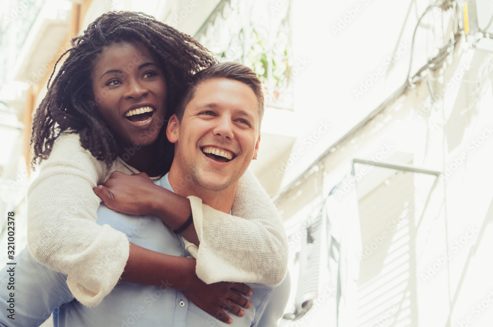 Young man laughing and carrying girlfriend on back outdoors. Happy interracial couple in street. Romance and happiness concept. Front view. - obrazy, fototapety, plakaty 