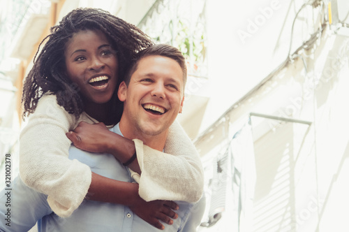 Young man laughing and carrying girlfriend on back outdoors. Happy interracial couple in street. Romance and happiness concept. Front view. photo