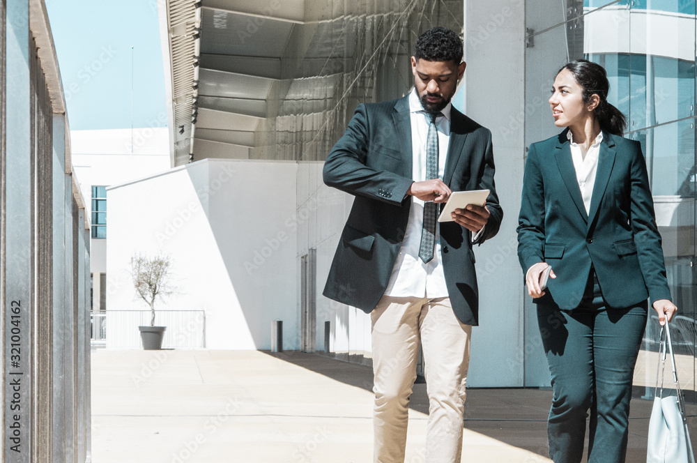 Business people talking and walking along street. Business man and woman wearing formal clothes and carrying gadgets with building in background. Business people concept. Front view. Business people talking and walking along street. Business man and woman wearing formal clothes and carrying gadgets with building in background. Business people concept. Front view.