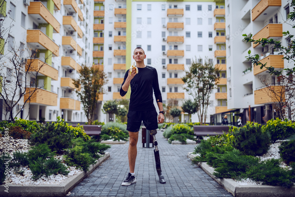 Full length of attractive caucasian sportsman with artificial leg standing in park surrounded by buildings and eating banana.