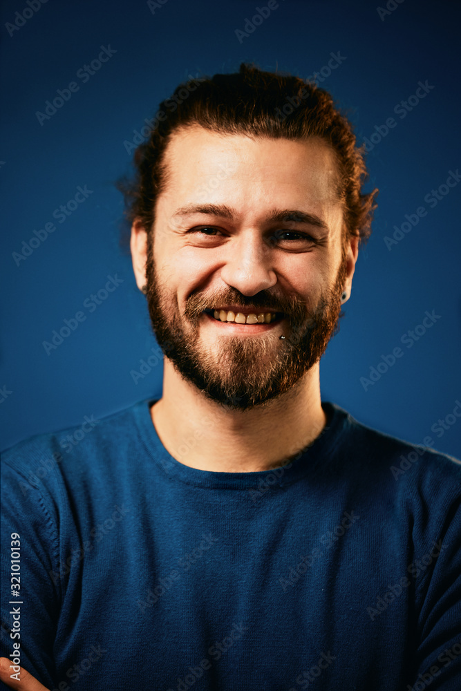 Young charming attractive Caucasian bearded hipster with curly hair dressed in sweater standing in front of blue background and looking at camera.