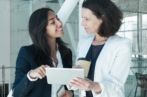 Two positive business women pointing at tablet screen in hall. Colleagues or friends standing with tablet computer. Technology and news concept.