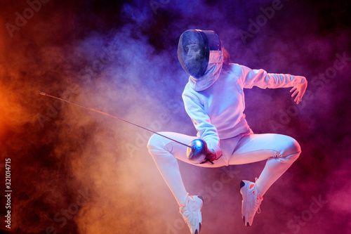 Blooming. Teen girl in fencing costume with sword in hand isolated on black background, neon lighted smoke. Practicing and training in motion, action. Copyspace. Sport, youth, healthy lifestyle.