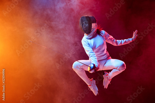 Fototapeta Naklejka Na Ścianę i Meble -  Blooming. Teen girl in fencing costume with sword in hand isolated on black background, neon lighted smoke. Practicing and training in motion, action. Copyspace. Sport, youth, healthy lifestyle.