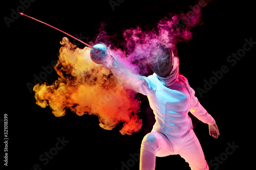 Contrasts. Teen girl in fencing costume with sword in hand isolated on black background, neon lighted smoke. Practicing and training in motion, action. Copyspace. Sport, youth, healthy lifestyle. photo