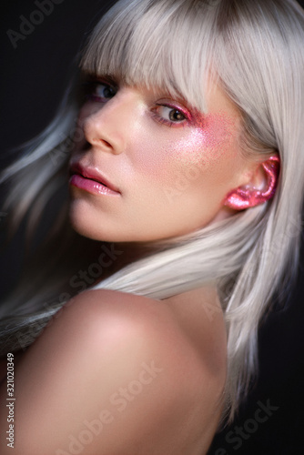 Portrait of attractive blonde woman with glittering eye and ear makeup looking at camera on dark studio background