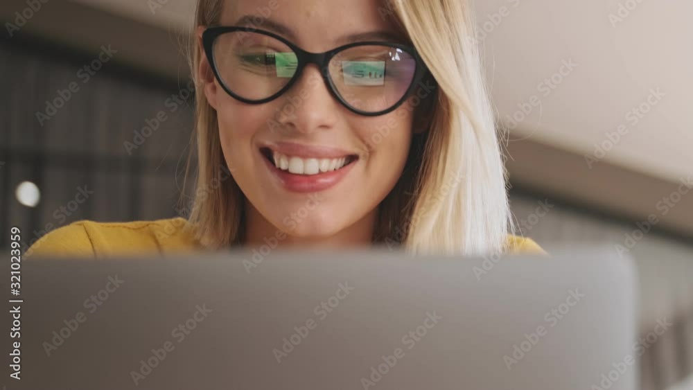 A close-up view of a smiling blonde woman wearing eyeglasses is working on her laptop while sitting in a conference hall