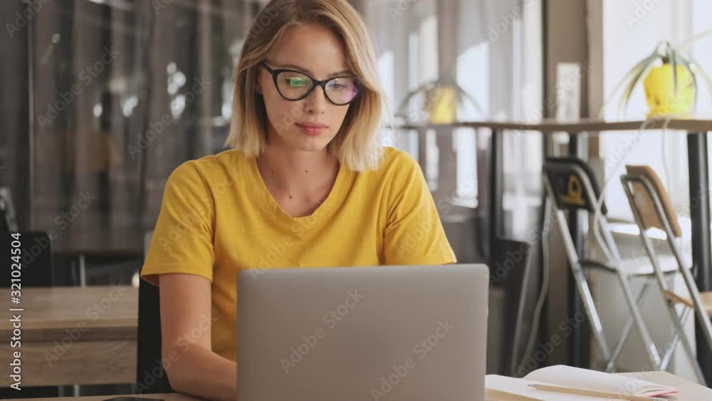 A concentrated blonde woman wearing eyeglasses and yellow t-shirt is working on her laptop while sitting in a conference hall