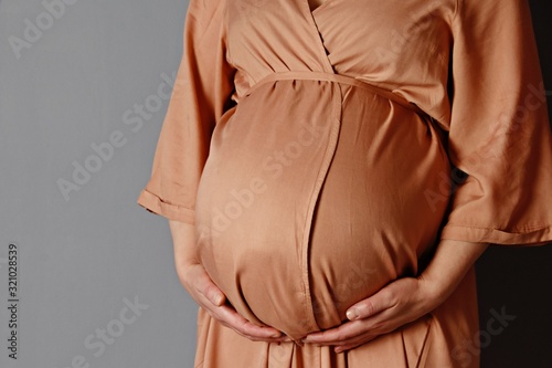 Beautiful pregnant woman touching her belly with hands on a grey background. Young mother anticipation of the baby. Image of pregnancy and maternity. Close-up, copy space, indoors.