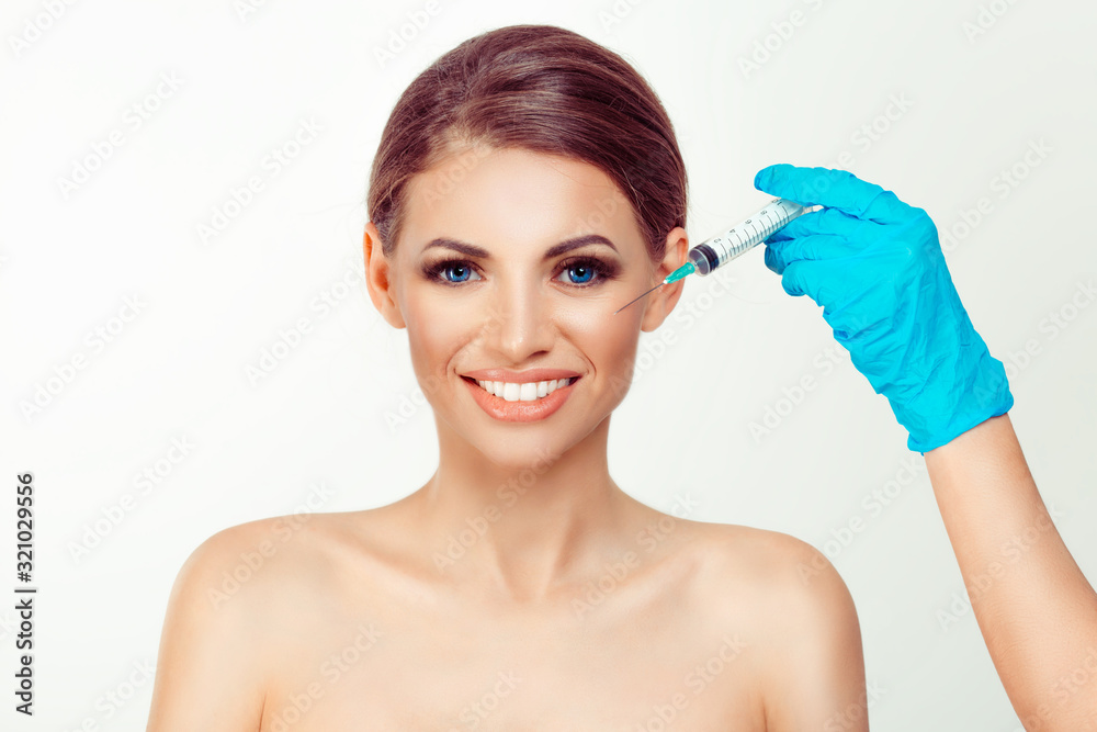 Smiling beautiful woman ready for anti aging serum shot in cheek doctor hand blue glove hold syringe over girl face Peaceful pretty model white background having botulinum toxin injection on the cheek