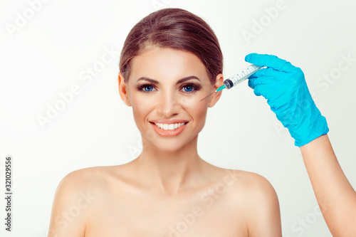 Smiling beautiful woman ready for anti aging serum shot in cheek doctor hand blue glove hold syringe over girl face Peaceful pretty model white background having botulinum toxin injection on the cheek