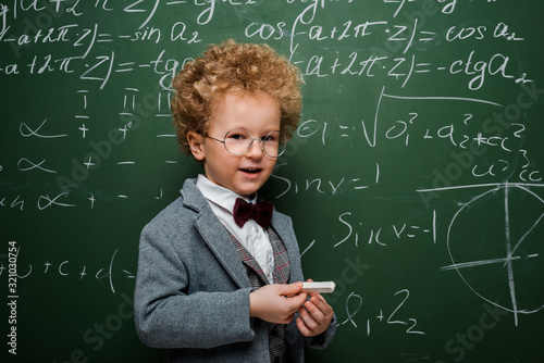 cute and smart child in suit with bow tie holding chalk near chalkboard with mathematical formulas