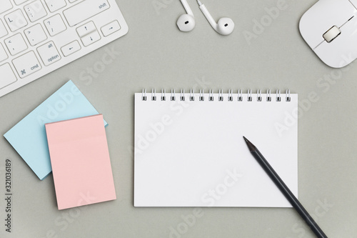 A blank white notebook for notes with a pencil on a gray background surrounded by a keyboard, headphones and mouse. Creative flat lay photo of workspace desk with copy space. 