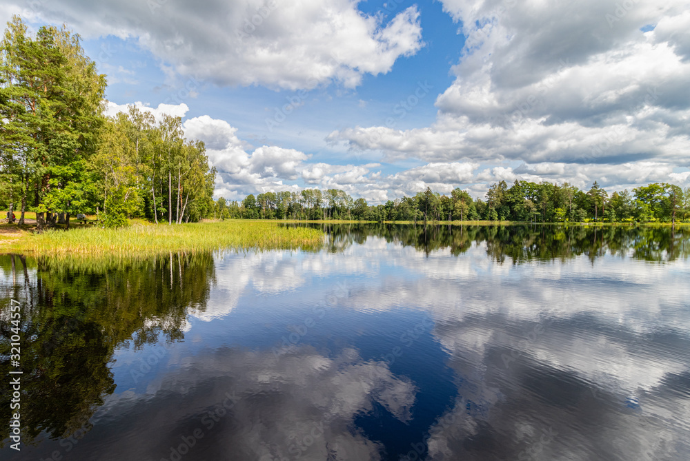 Wonderful landscape with lake on a sunny summer day. Blue sky with cumulus clouds, forest on the other side reflected in calm water. Latvia.