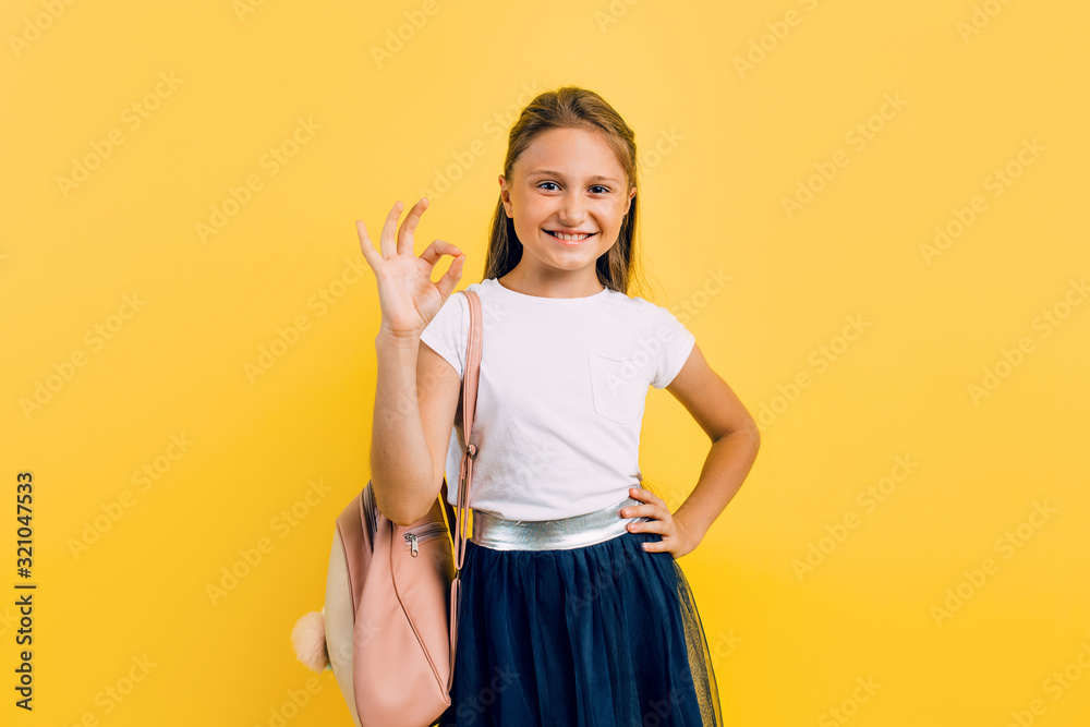 A little schoolgirl makes an approving gesture with her hand, OK on a yellow background. A little schoolgirl makes an approving gesture with her hand, OK on a yellow background.