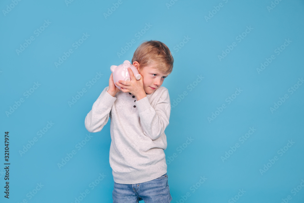 Cute little boy shaking piggy box on blue background Cute little boy shaking piggy box on blue background