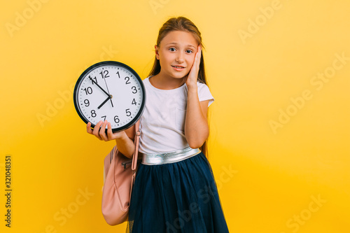 A teenage girl with a watch in her hands looks at the clock on a yellow background in fear