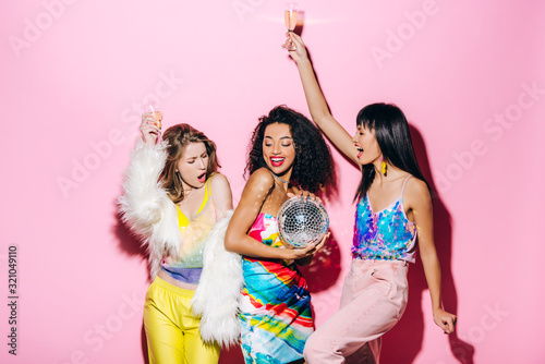 excited multicultural girlfriends with champagne and disco ball having fun on pink photo
