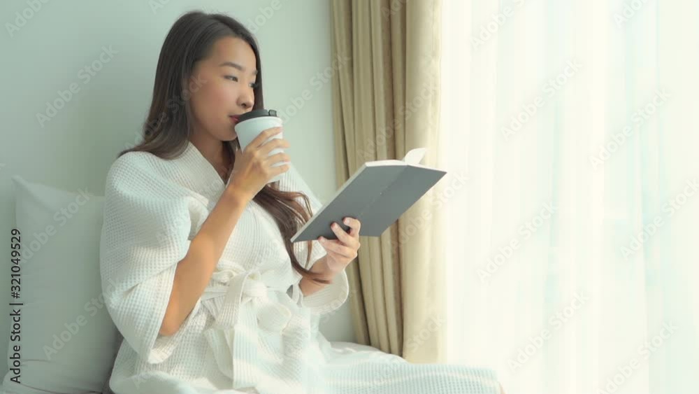 Young asian woman drinking morning coffee and reading an interesting book or an article while sitting on a bed near a window and wearing a white dressing gown. Young asian woman drinking morning coffee and reading an interesting book or an article while sitting on a bed near a window and wearing a white dressing gown.