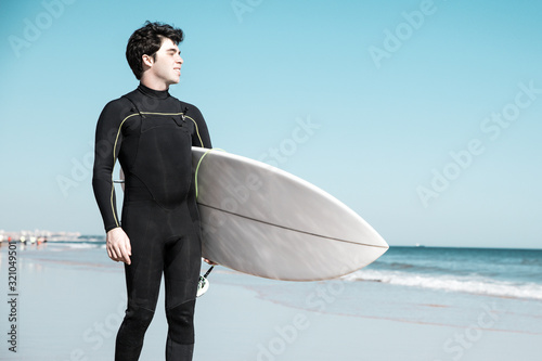 Smiling handsome young man holding surfboard on sunny beach. Handsome guy wearing wetsuit and standing. Surfboarding concept.