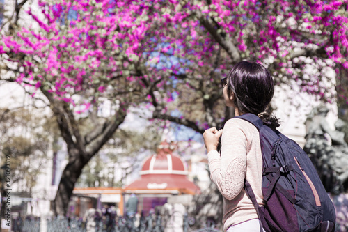 Woman wearing backpack and walking around town. Pretty young lady looking at city sights with blossoming spring tree in background. Tourism concept.