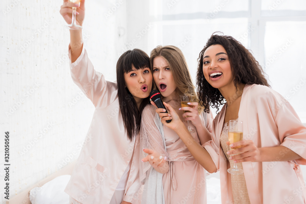 excited multiethnic girls holding champagne and singing with microphone on pajama party excited multiethnic girls holding champagne and singing with microphone on pajama party